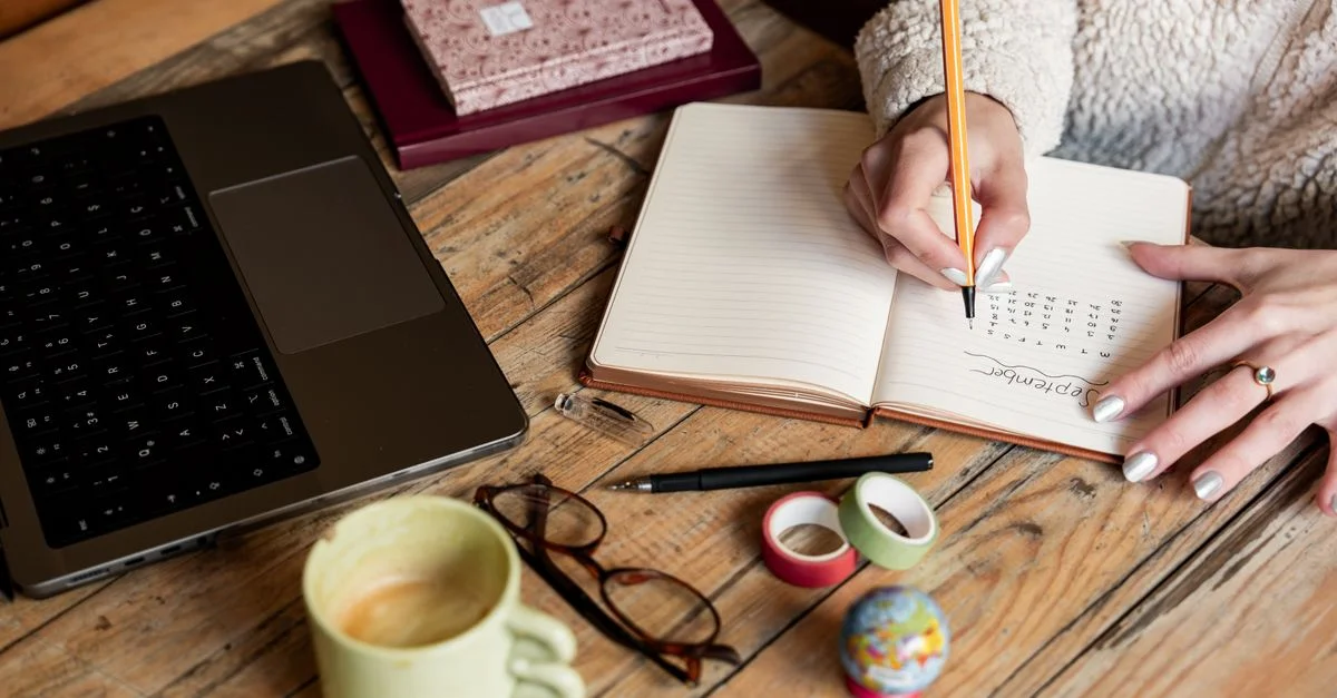 Une femme planifiant son emploi du temps dans un carnet à côté d'un ordinateur portable et d'un café sur un bureau en bois.