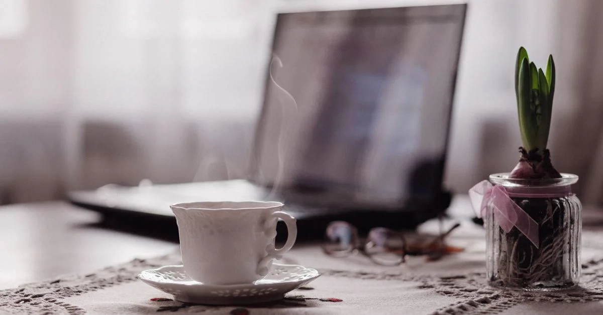 Un espace de travail intérieur serein avec une tasse de thé fumante, un ordinateur portable et un bulbe de printemps en pot, le tout sur une nappe décorative.