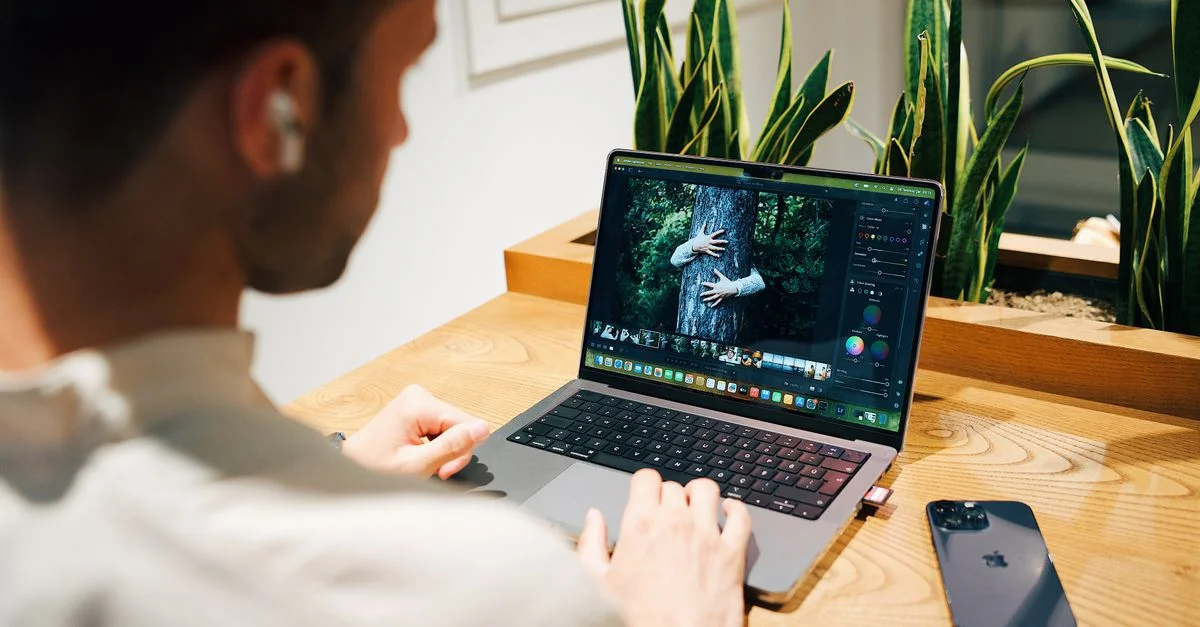 Un homme utilisant un ordinateur portable pour le retouche photo dans un environnement de bureau moderne, avec des plantes et un smartphone à proximité.