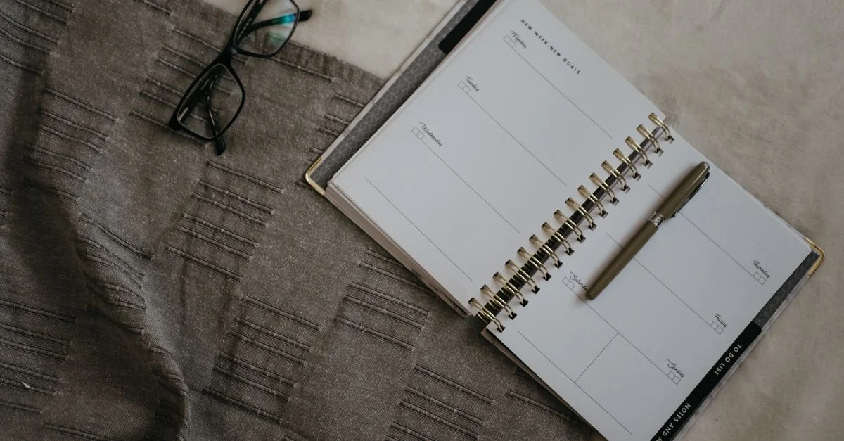 Une image en flatlay d'un planificateur, de lunettes et d'un stylo sur une surface en tissu texturé.